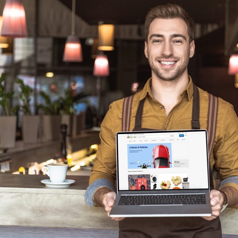 smiling young waiter holding laptop with ebay website on screen in cafe
